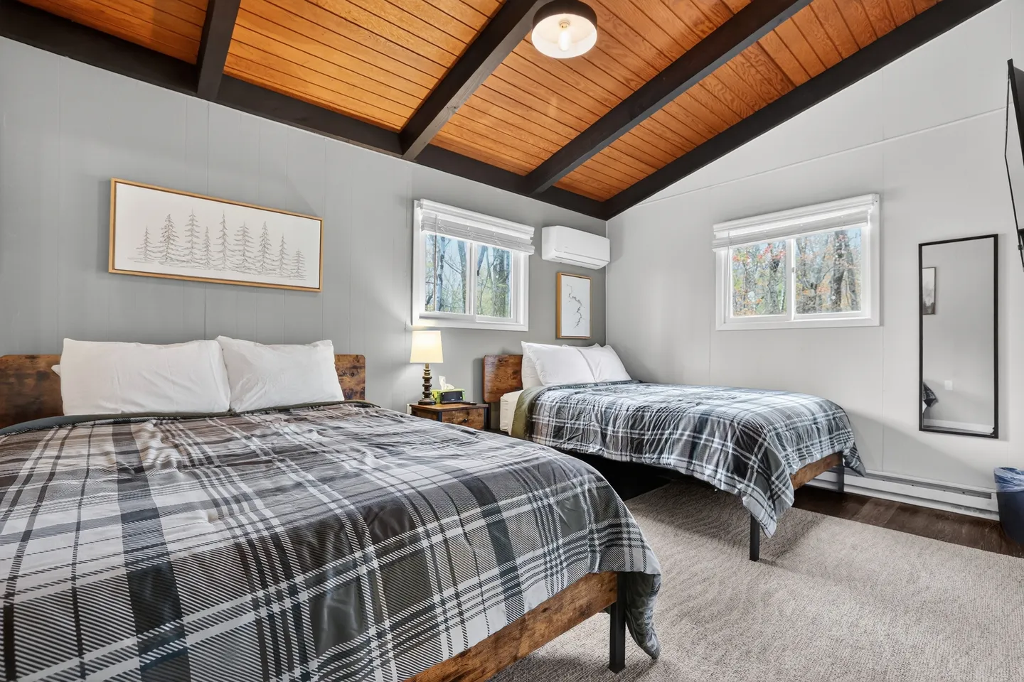 Bedroom with two beds topped with plaid quilts, a wooden sloped ceiling with dark beams, and two windows.