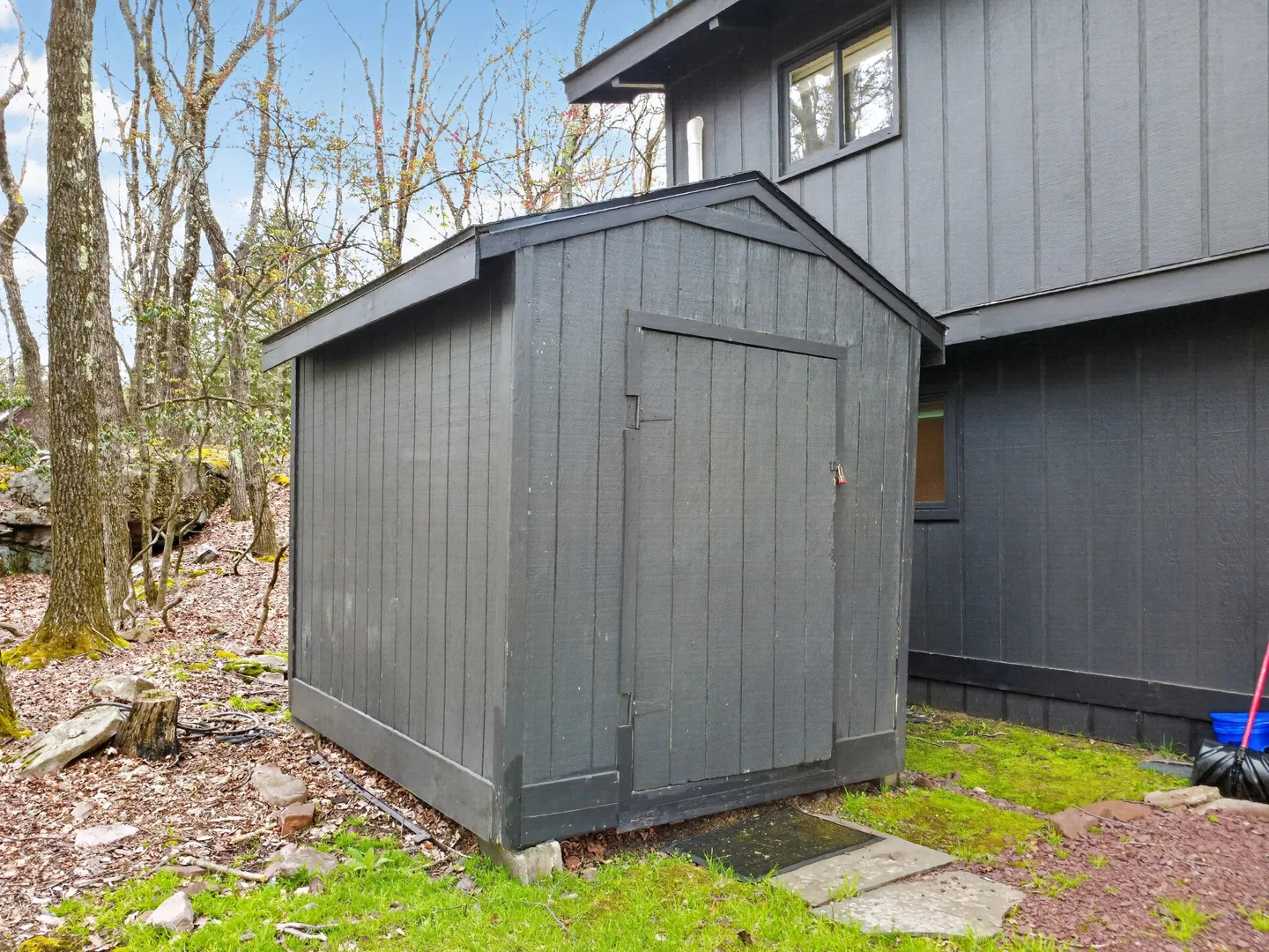 Exterior dark gray storage shed standing near the side of a matching house surrounded by trees.