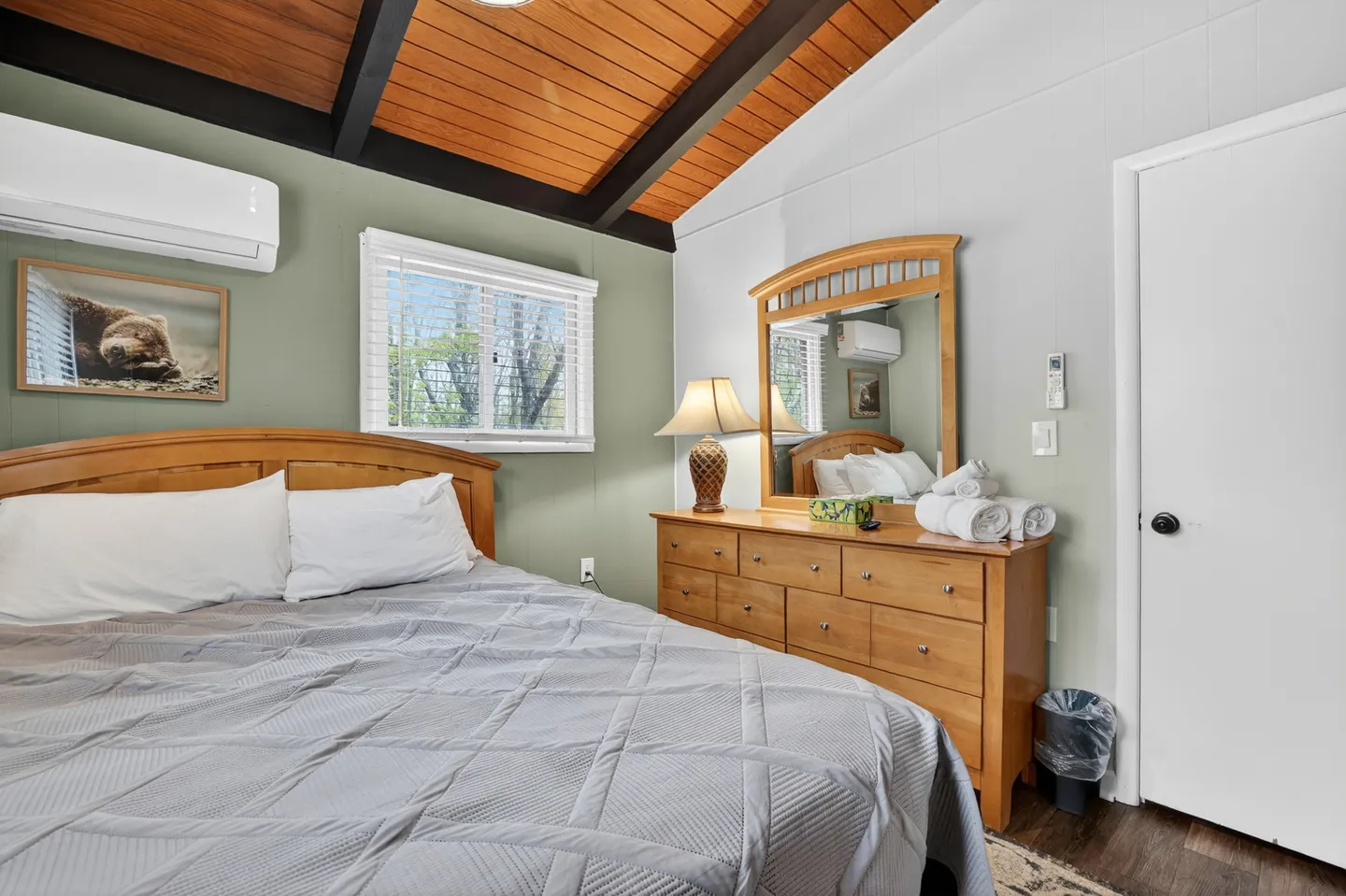 Bedroom with vaulted wood-paneled ceiling, a made bed with a light gray quilt, a wooden dresser with a mirror, and a wall-mounted AC unit.