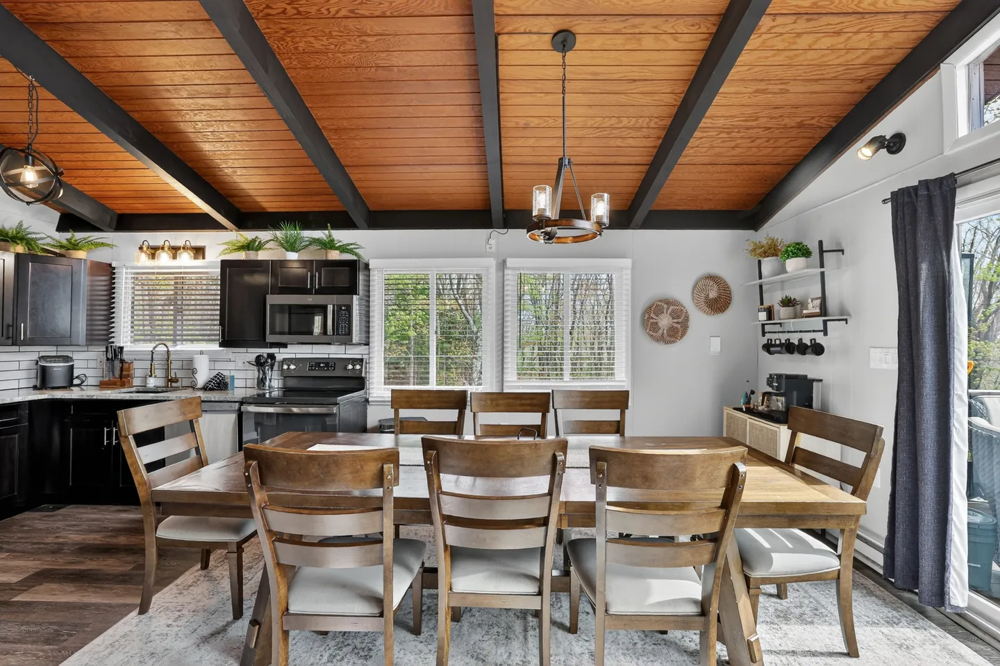 Dining area with a large wooden table and chairs beneath a wood-paneled ceiling with dark beams, adjacent to an open kitchen.