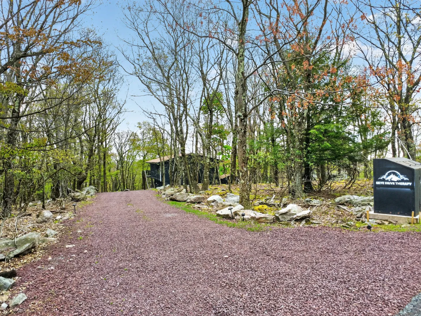 A gravel driveway lined with rocks and trees leading toward a cabin in the woods with a property sign on the right.