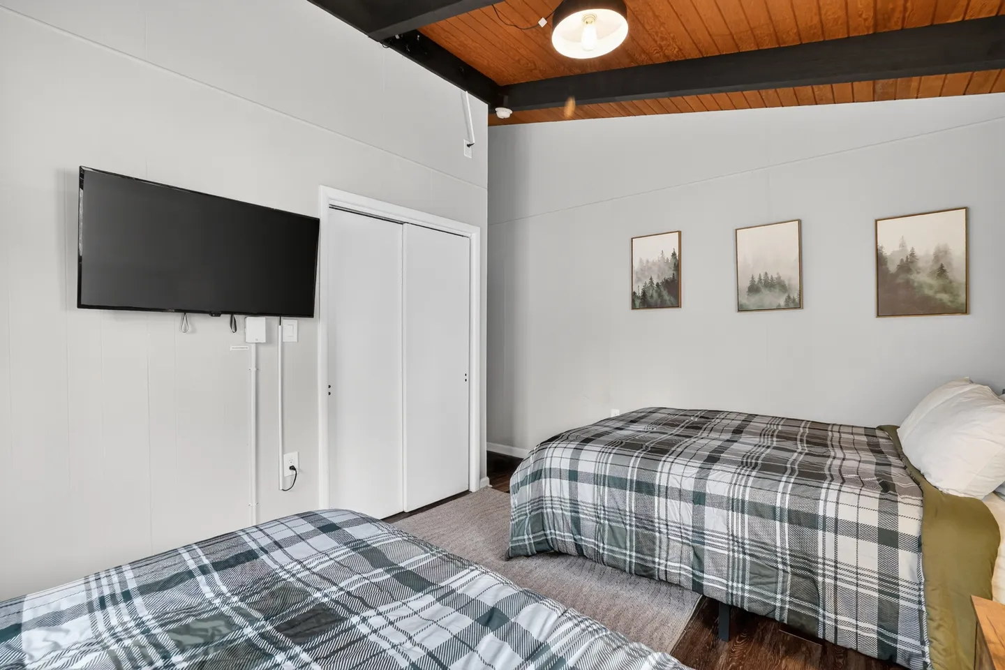 Bedroom featuring two beds with plaid bedding, a wall-mounted flat-screen TV, and an angled wood-paneled ceiling with dark exposed beams.
