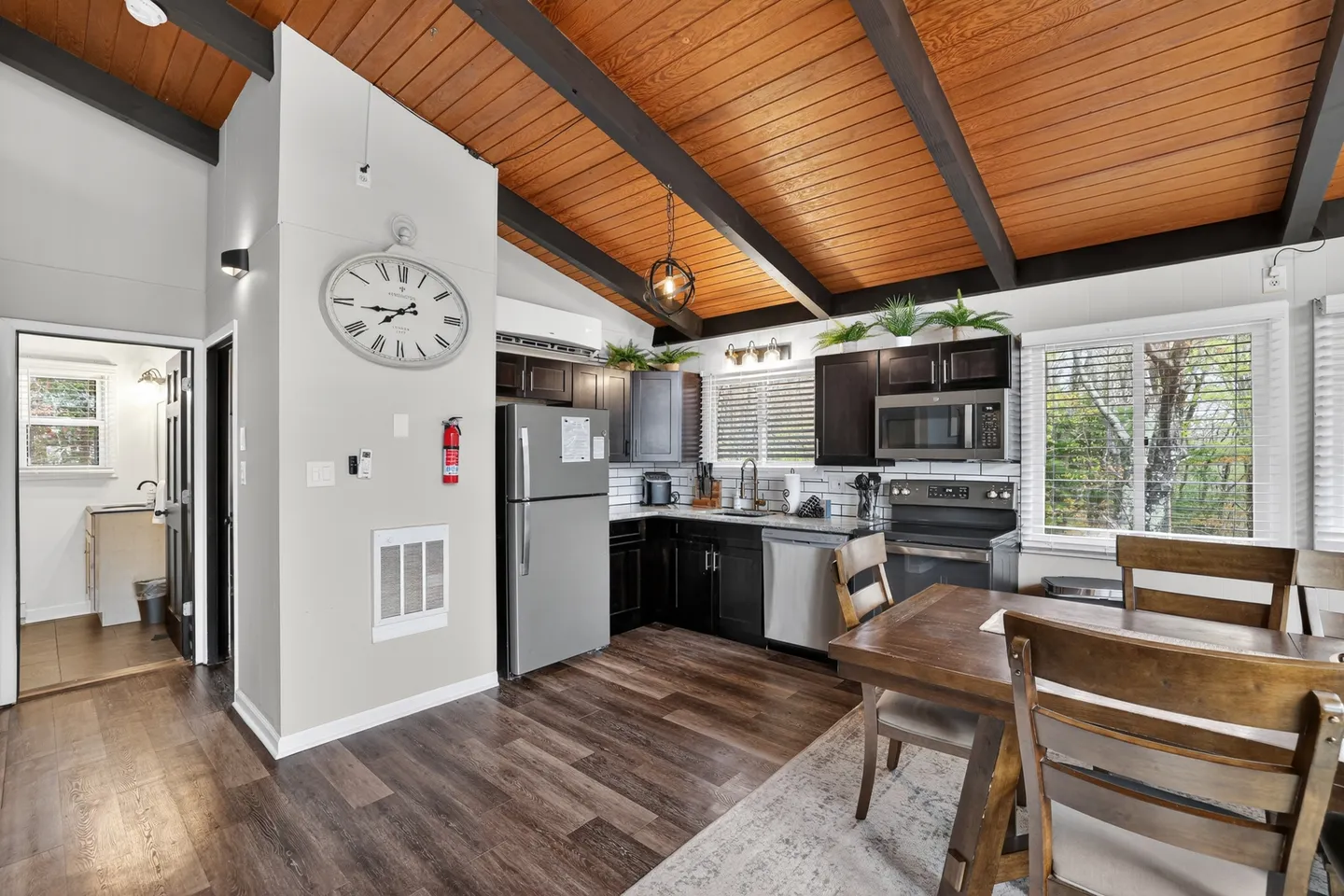 Open dining and kitchen area featuring dark wood cabinets, stainless steel appliances, wood-paneled vaulted ceiling with dark beams, and a wooden dining table.