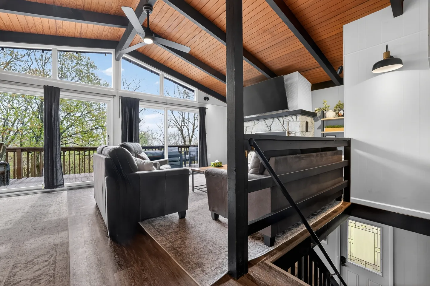 Living room with vaulted wood-paneled ceiling, dark beams, upholstered seating, and floor-to-ceiling windows leading to a deck.