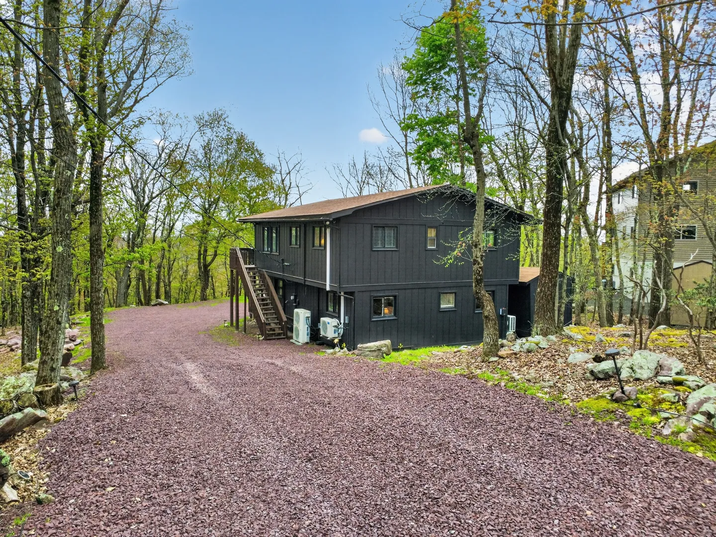 Exterior of a two-story dark gray house nestled among trees with a gravel driveway in front.