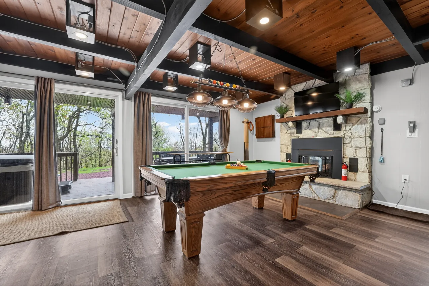 Game room featuring a wooden pool table centered below modern hanging lights, a stone fireplace, and sliding glass doors to a deck.