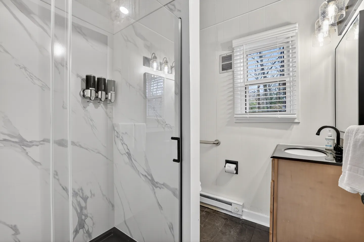 Bathroom showing a walk-in glass shower with marble-patterned tile, next to a vanity and a window with blinds.