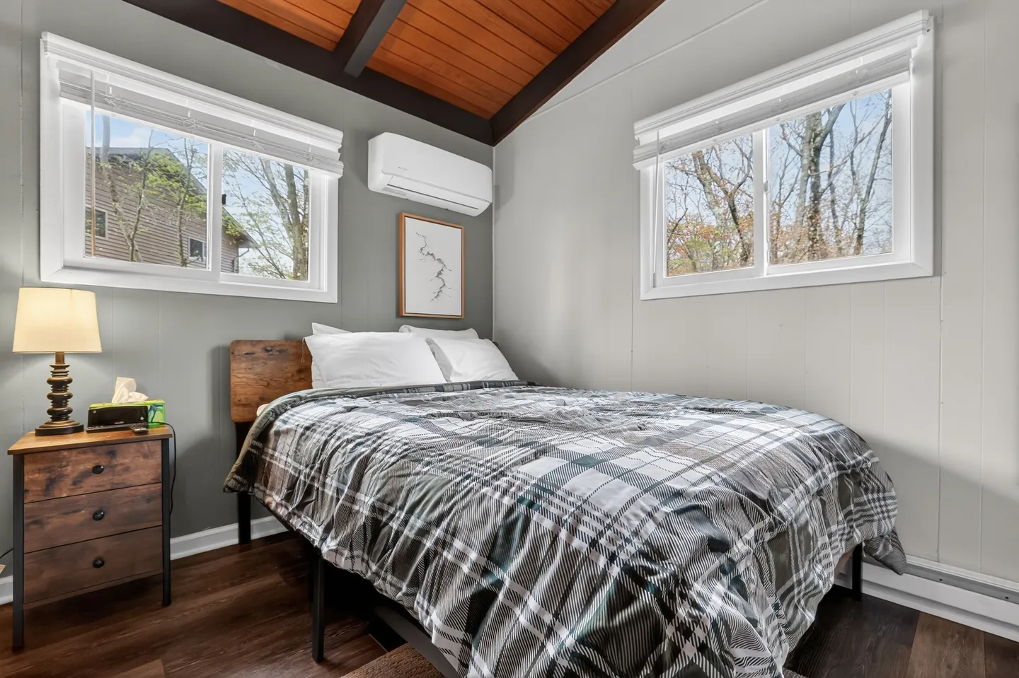 Bedroom with a plaid-covered bed, wood-paneled ceiling, two windows, nightstand with a lamp, and a wall-mounted mini-split unit.