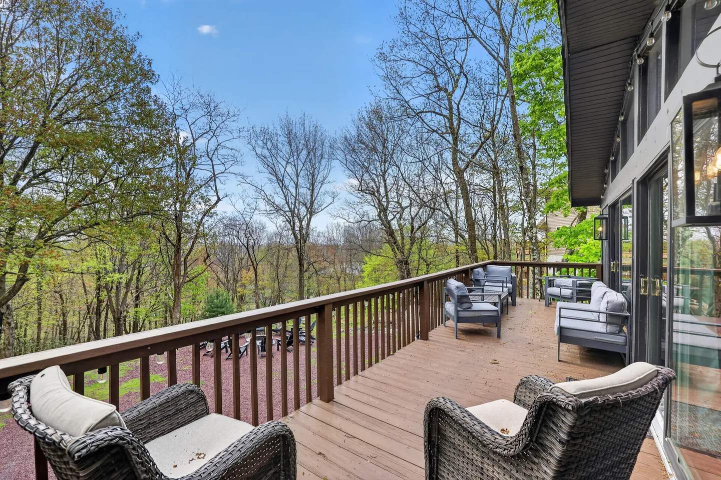 Elevated wooden deck with outdoor armchairs overlooking a wooded yard and a lower fire pit area under a blue sky.