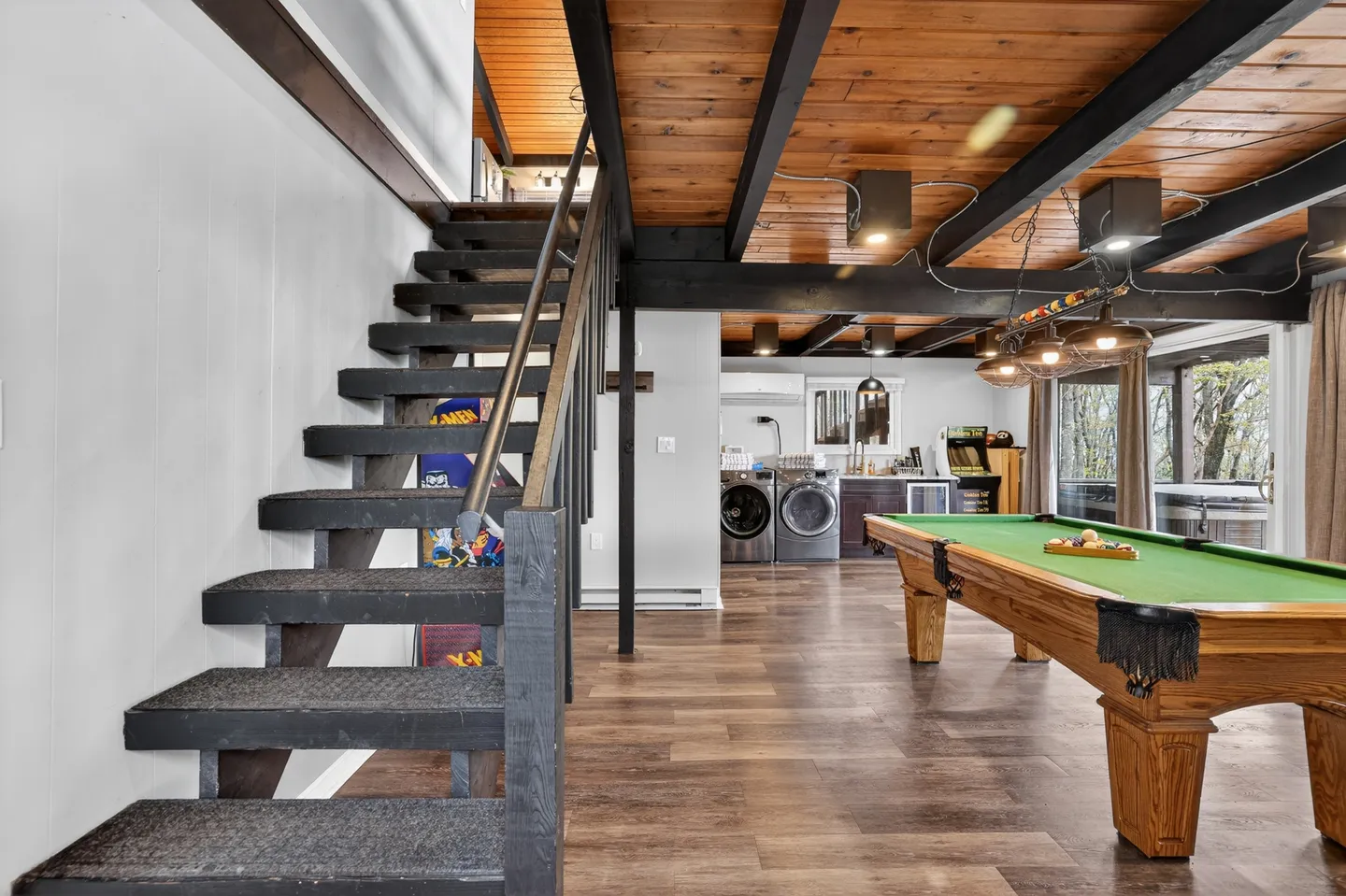 A game room with an open wooden staircase leading to the upper floor, a green-felt pool table, and a laundry area in the background.