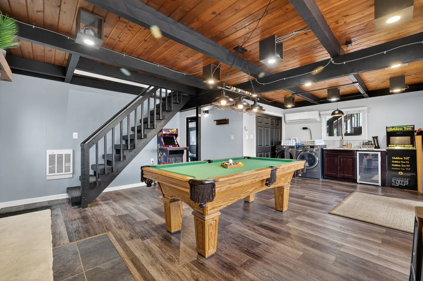 Game room featuring a green-felt pool table, arcade machines, stairs, and a laundry area beneath a wood-beamed ceiling.