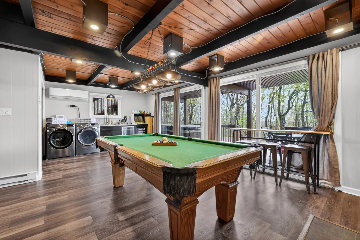Game room featuring a wooden pool table with green felt, dark ceiling beams with wood paneling, sliding glass doors to a deck, and laundry appliances in the corner.
