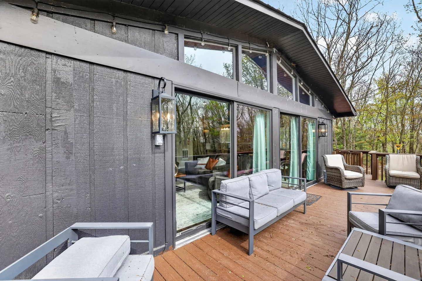 Outdoor wooden deck with modern patio furniture outside a dark-paneled house with large glass sliding doors.