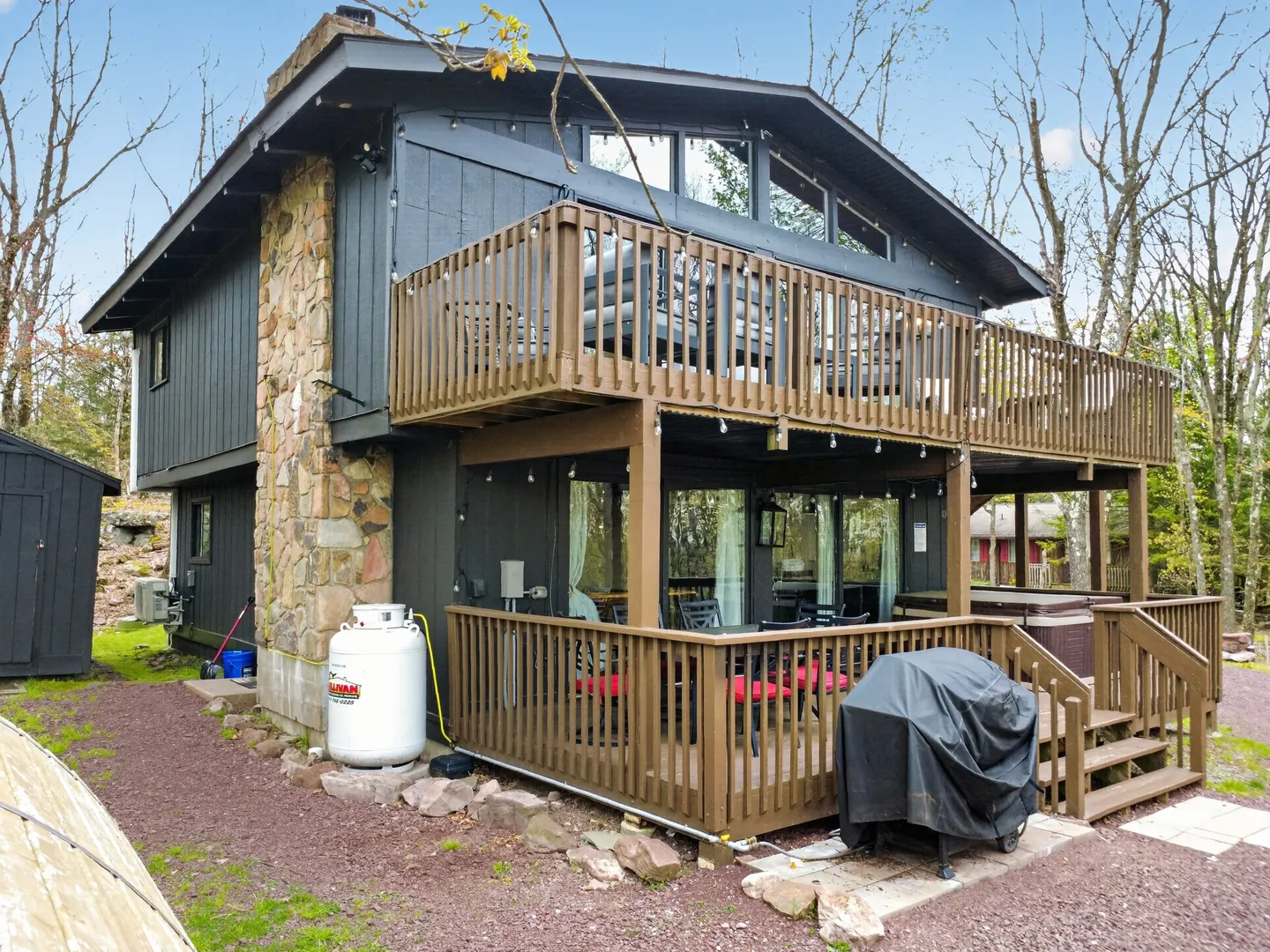 Exterior rear view of a two-story dark siding home with stone chimney, upper balcony, and lower covered wooden deck with grill and hot tub.