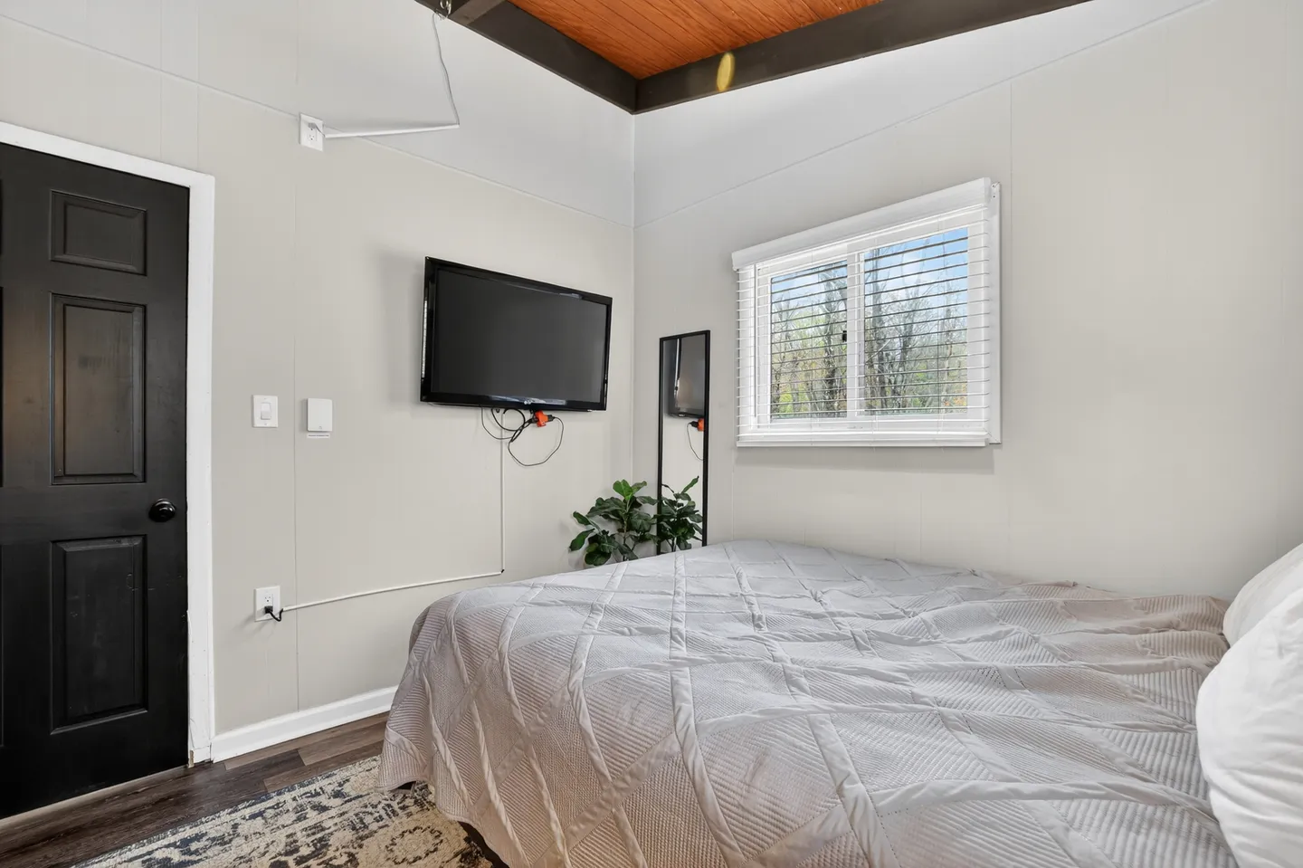 Bedroom with a bed made with a light gray quilt, a wall-mounted TV, a dark door, and a window with white blinds.