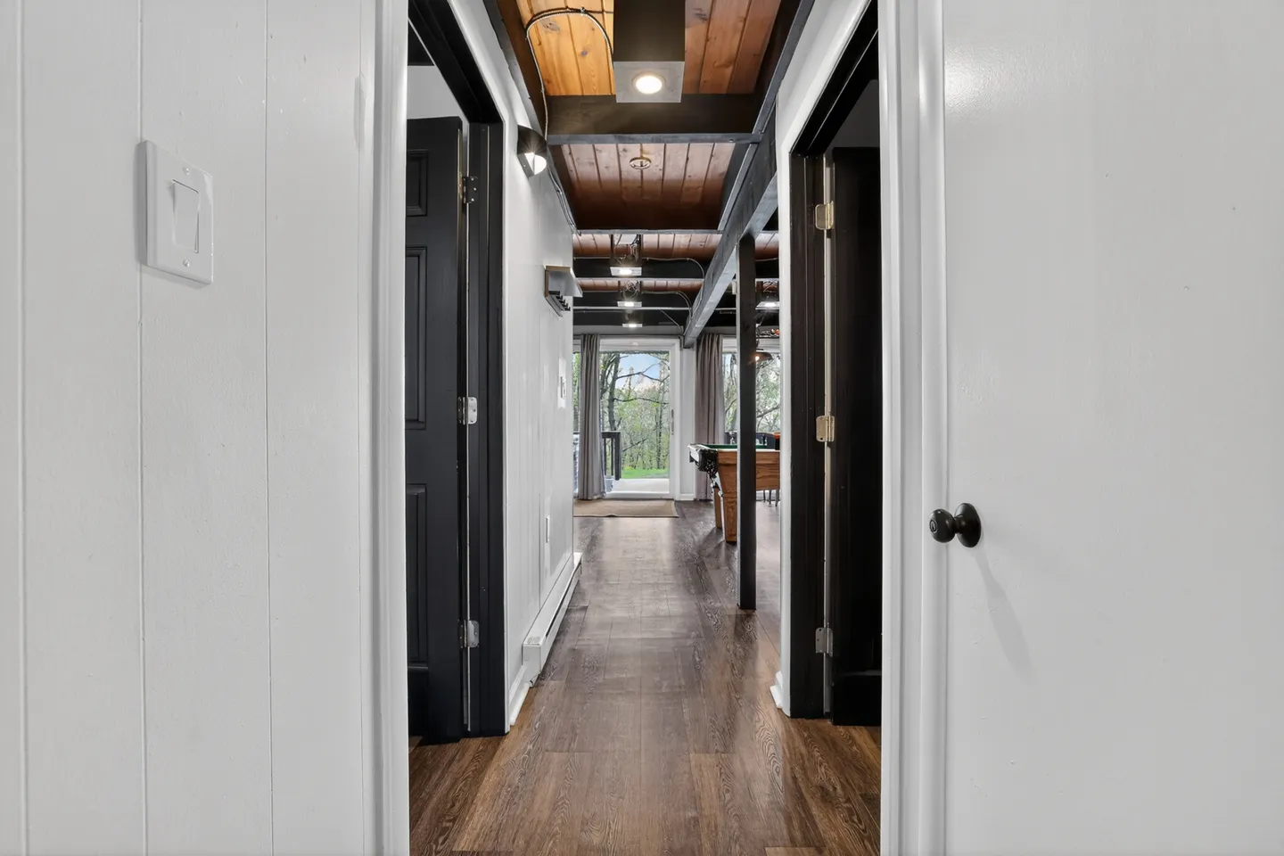 A hallway viewed through a doorway, featuring wood flooring, black doors, and an exposed wood-paneled ceiling with beams leading to a recreation room with a pool table.