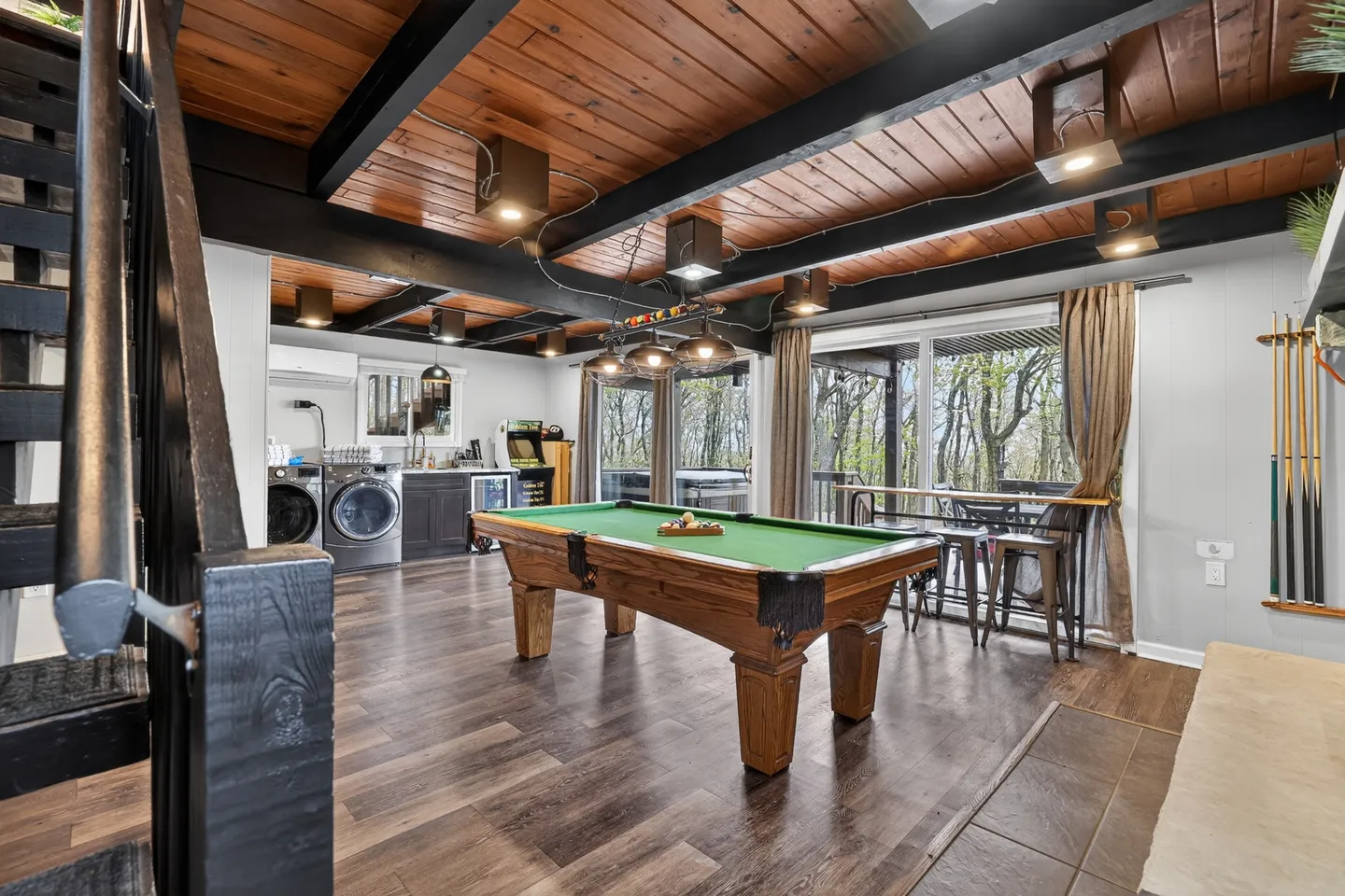 Game room featuring a green-felt pool table beneath a wood-paneled ceiling with exposed black beams, alongside a laundry and bar area and sliding glass doors.