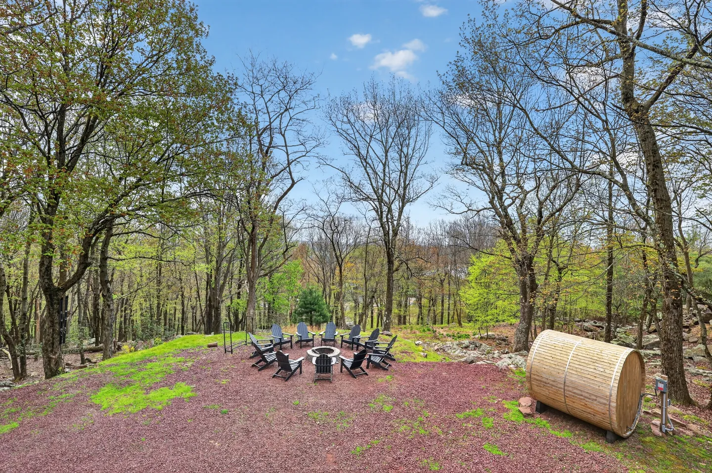 Outdoor clearing with a circular fire pit surrounded by Adirondack chairs, a barrel sauna to the right, and tall trees in the background under a blue sky.