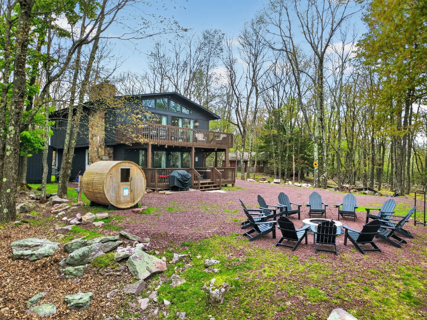 Exterior backyard view of a two-story dark cabin featuring a barrel sauna, upper and lower decks, and an outdoor fire pit with Adirondack chairs surrounded by trees.