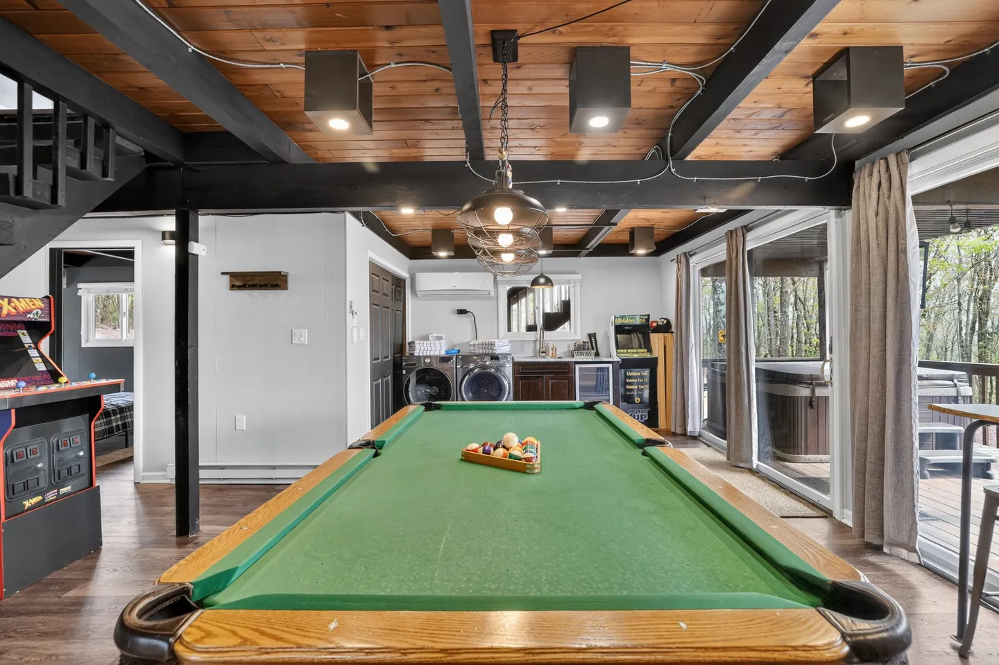 Game room featuring a green felt pool table in the center, arcade games, exposed beam ceiling, and sliding glass doors leading to an outdoor deck.