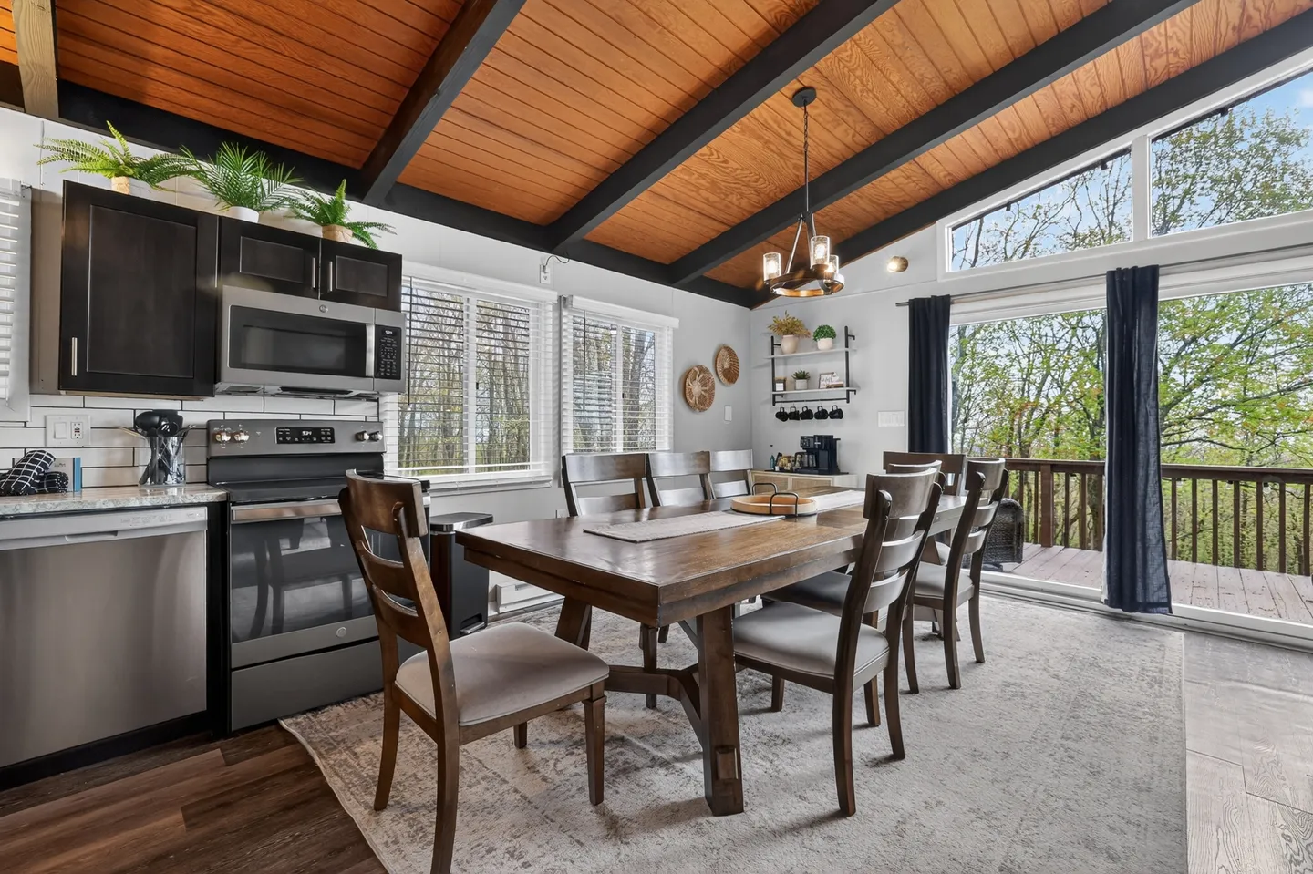 Dining area with a wooden table and chairs adjacent to a kitchen with stainless steel appliances under a vaulted wood-paneled ceiling.
