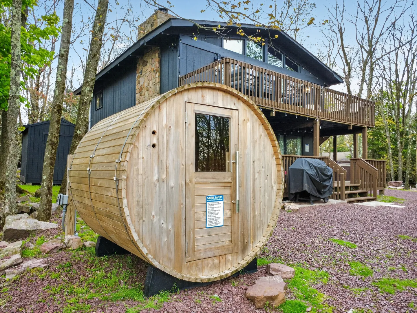 Outdoor wooden barrel sauna sitting on gravel ground in front of a multi-story cabin with a balcony.