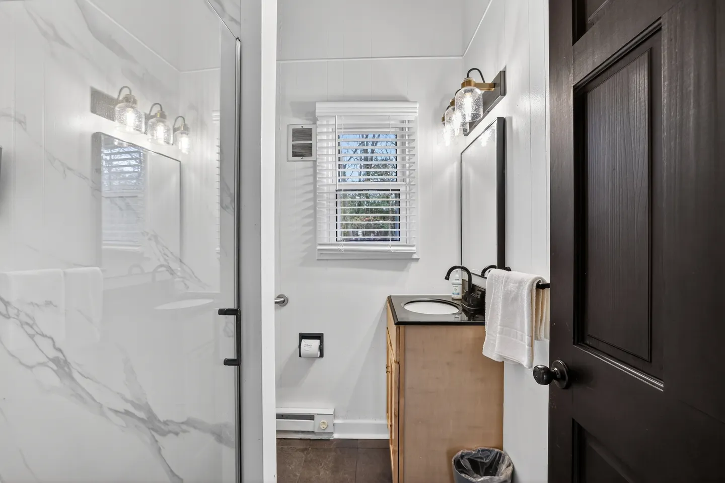 Bathroom featuring a vanity with a dark countertop, a mirror with an overhead vanity light, a window with white blinds, and a glass-enclosed shower with marble-look tile.