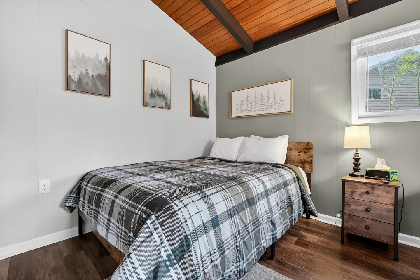 Bedroom with vaulted wood-plank ceiling, exposed dark beams, a plaid-covered bed, and framed forest artwork on grey walls.