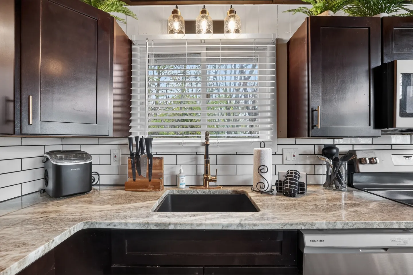 Kitchen sink area with dark cabinetry, light-toned countertops, white subway tile backsplash, and a window with blinds.