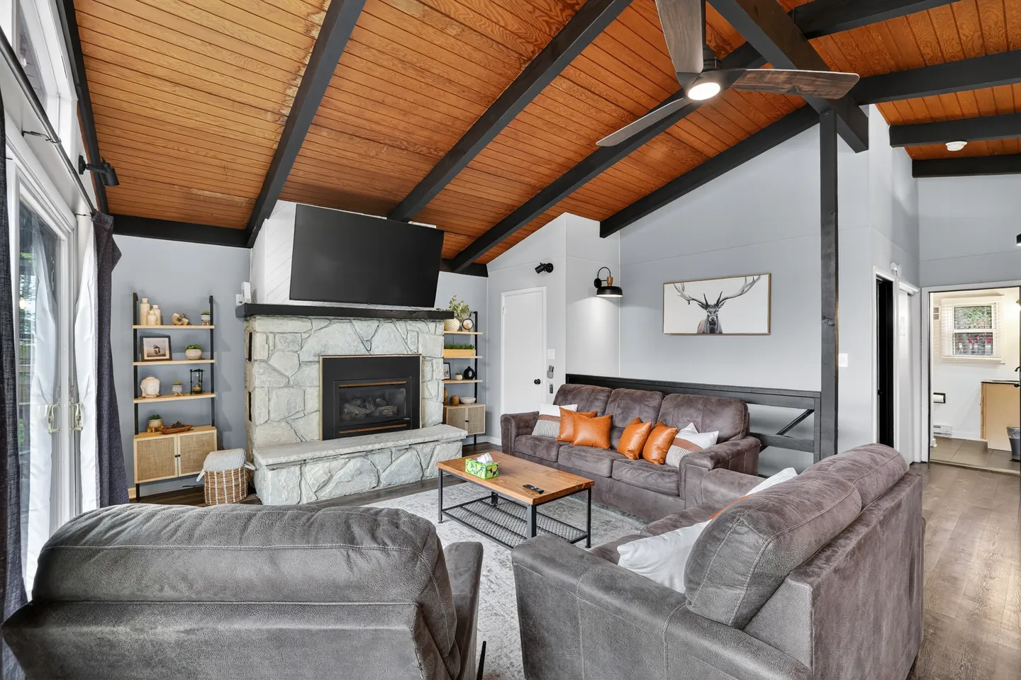 Living room with vaulted wood-paneled ceiling, dark exposed beams, a stone fireplace with a mounted TV, and gray leather sofas.