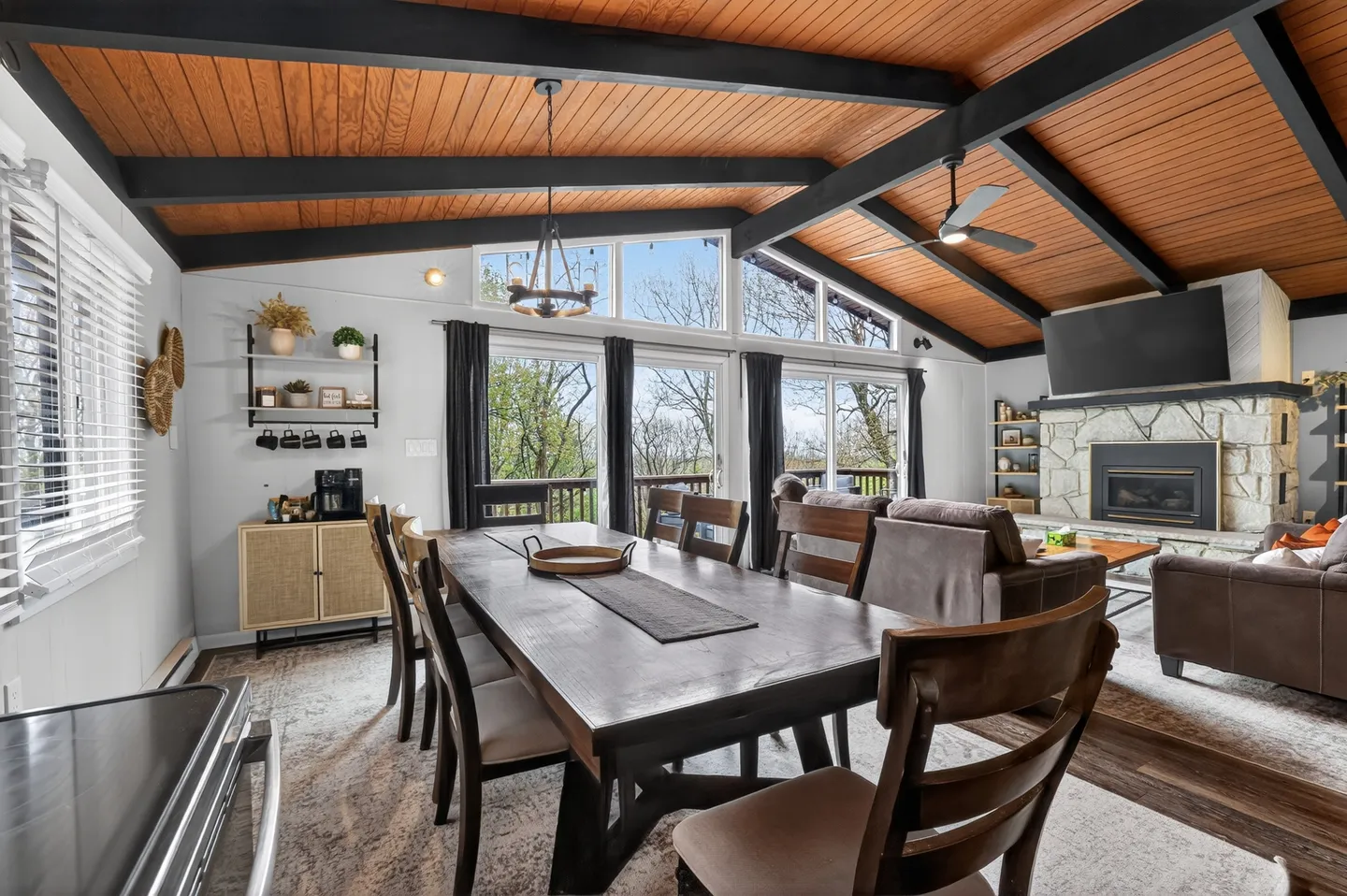 Open-concept dining and living area with wood-plank vaulted ceiling, exposed dark beams, dining table, and stone fireplace.