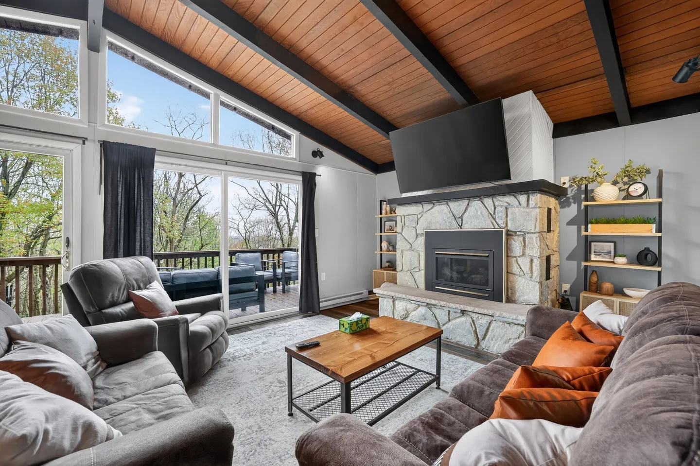 Living room with vaulted wood-paneled ceiling, stone fireplace with mounted TV, plush seating, and sliding glass doors leading to an outdoor deck.