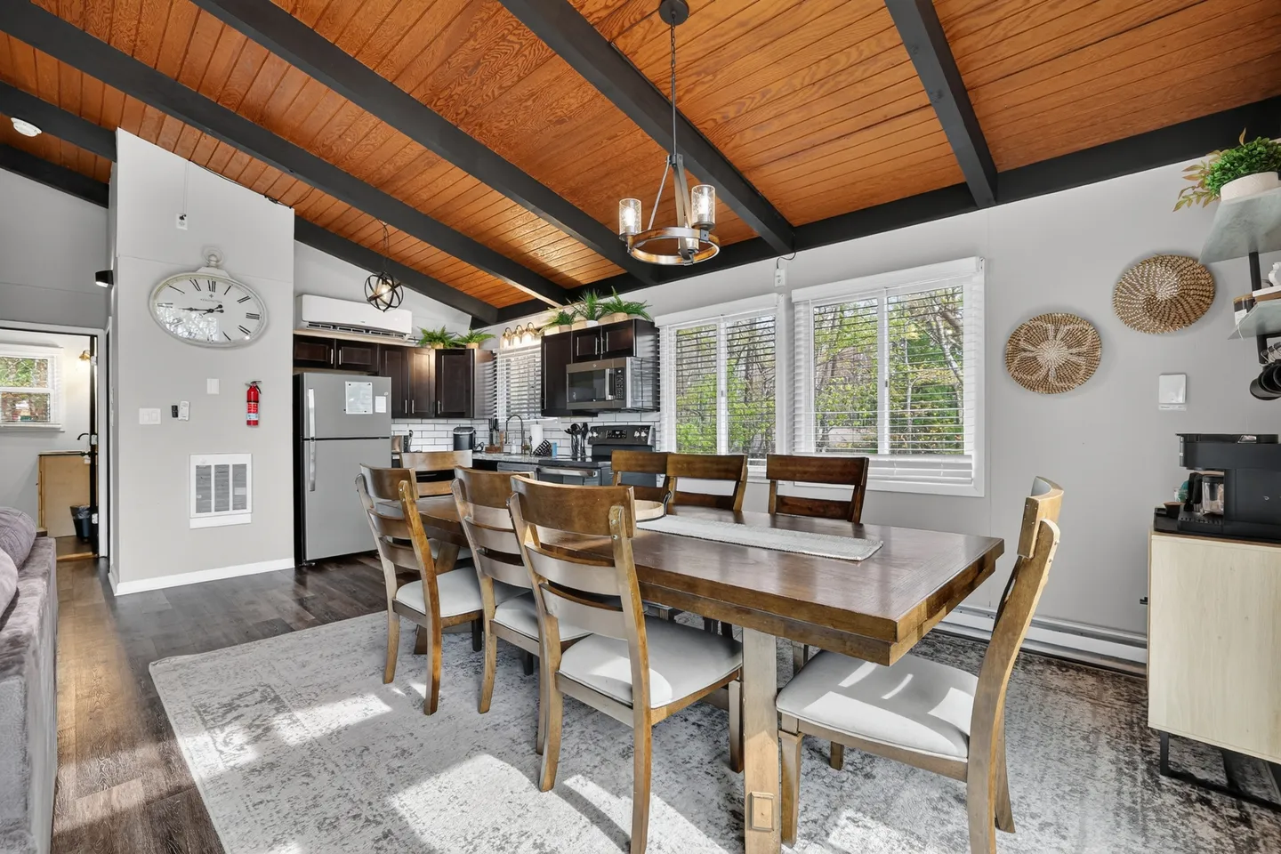 Dining area with a large wooden table and chairs, situated under a sloped wood-plank ceiling with dark exposed beams next to an open kitchen.