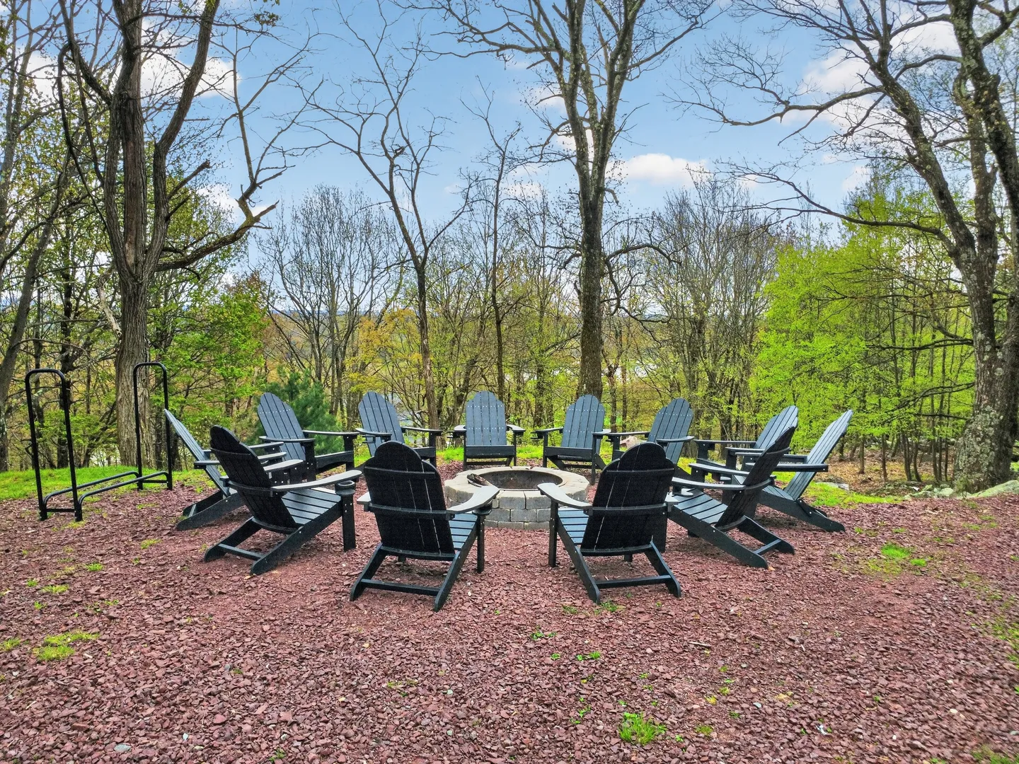Outdoor stone fire pit surrounded by a circle of dark Adirondack chairs on a reddish gravel clearing in a wooded area.