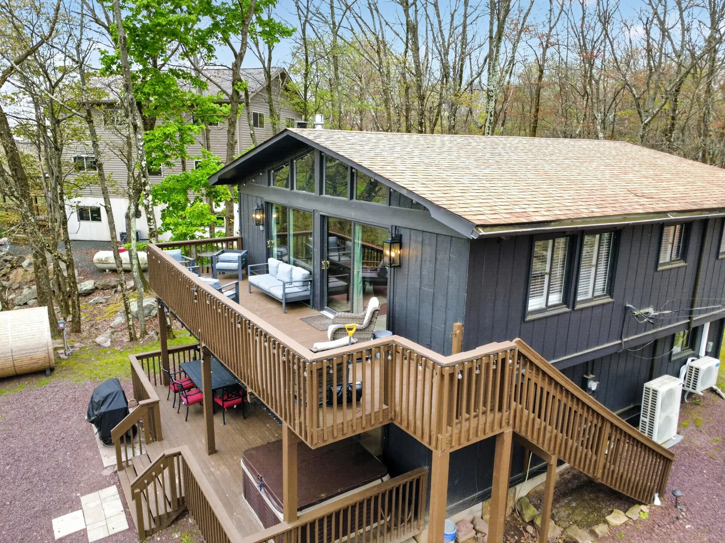Exterior elevated view of a dark-sided two-story cabin featuring multi-level wooden decks with outdoor furniture and a hot tub, surrounded by trees.