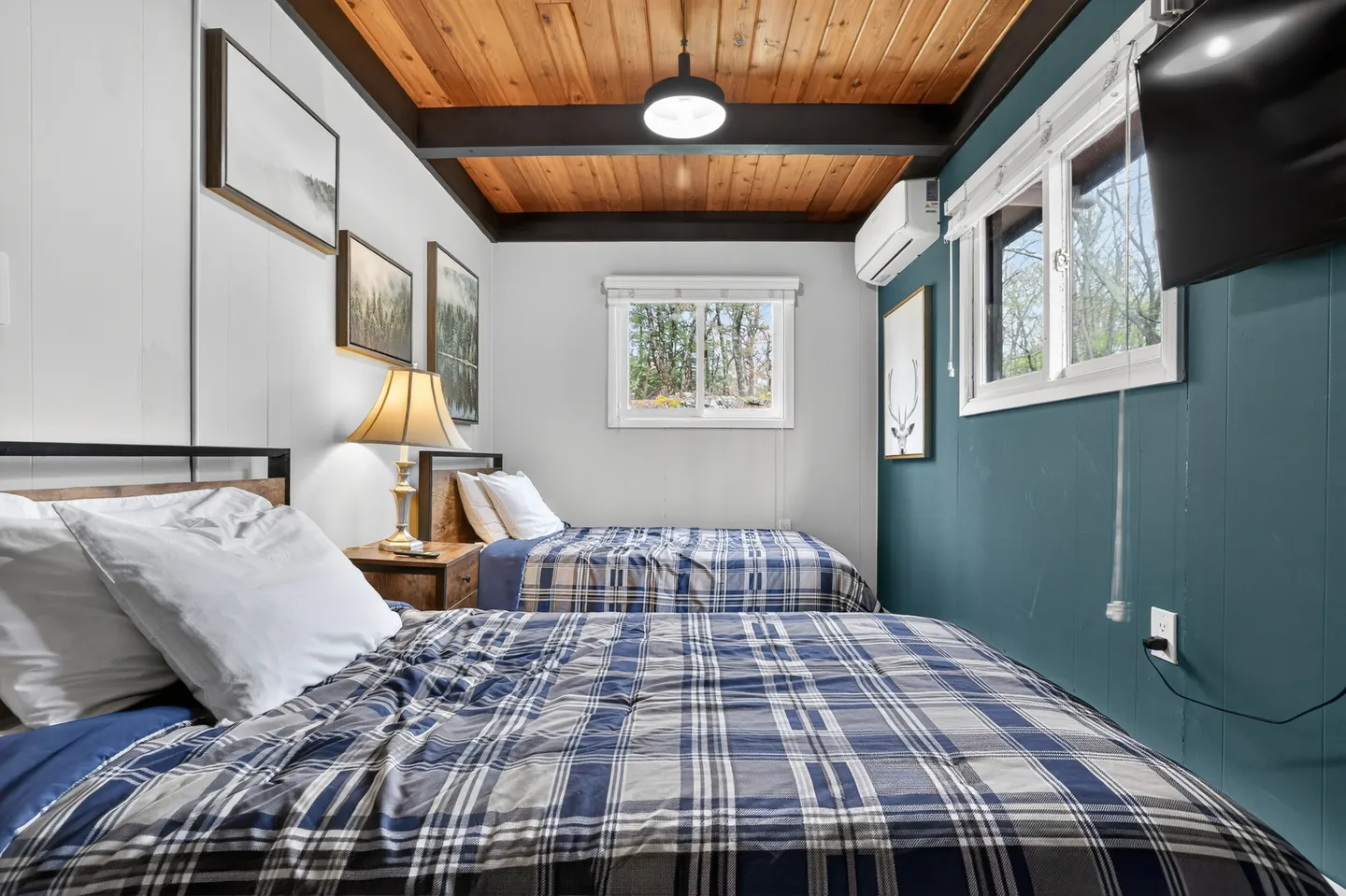 Bedroom with two beds dressed in blue and white plaid bedding, wood-plank ceiling, and teal accent wall.