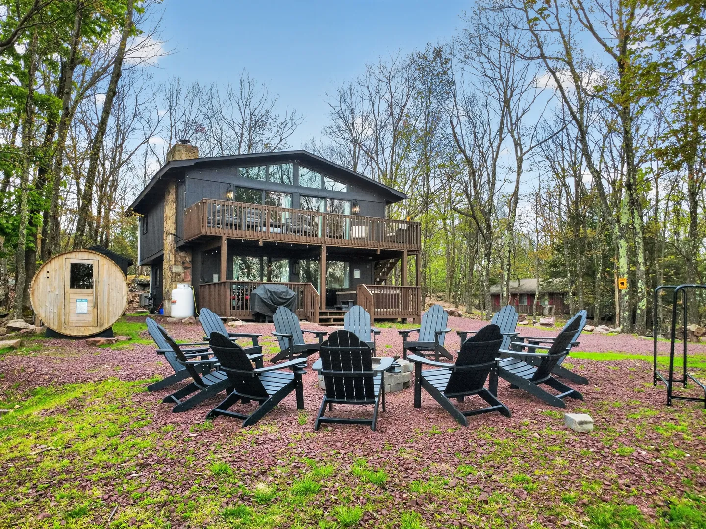 Exterior of a two-story home with upper and lower decks, a circular arrangement of Adirondack chairs around a fire pit, and a barrel sauna in the wooded yard.