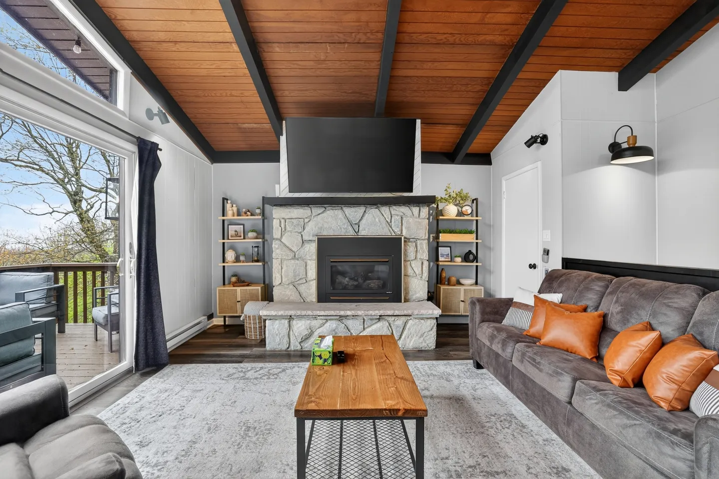 Living room with a stone fireplace, mounted TV, vaulted wood plank ceiling, sofa, and sliding glass door leading to an outdoor deck.