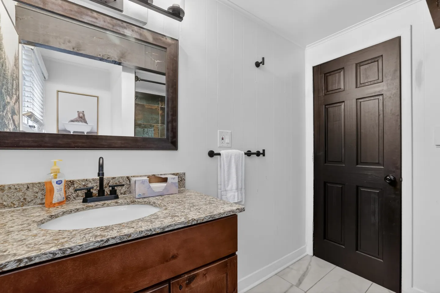 Bathroom vanity with a dark wood cabinet, speckled countertop, and a wood-framed mirror next to a dark paneled door.