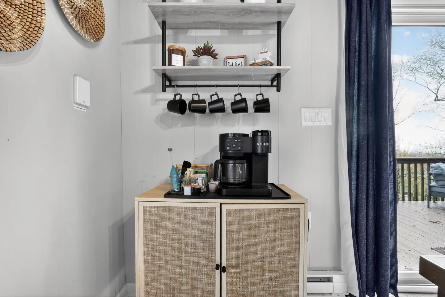 Dedicated coffee station featuring a cabinet, dual coffee maker, and wall-mounted shelves with hanging black mugs next to a sliding glass door.