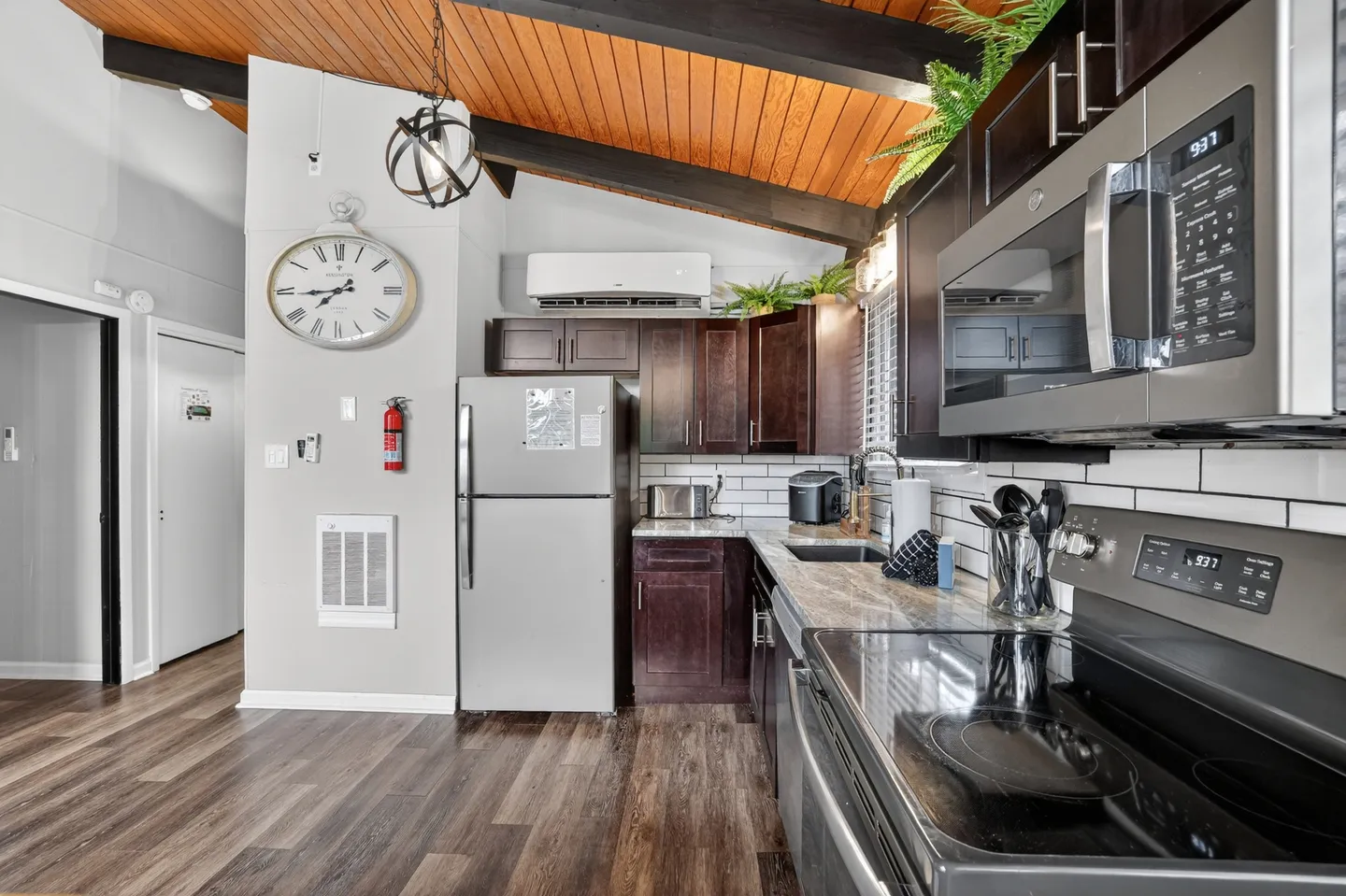 Kitchen featuring dark wood cabinetry, stainless steel appliances, wood-look flooring, and a slanted wood-paneled ceiling with exposed beams.
