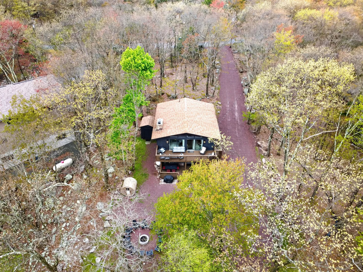 Aerial view of a cabin surrounded by woods with a deck, patio, and circular fire pit area.
