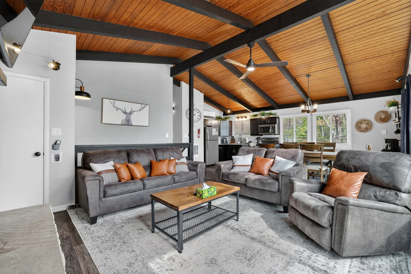 Living room with vaulted wood-plank ceiling, exposed beams, gray upholstered seating, and a view into the kitchen and dining area.