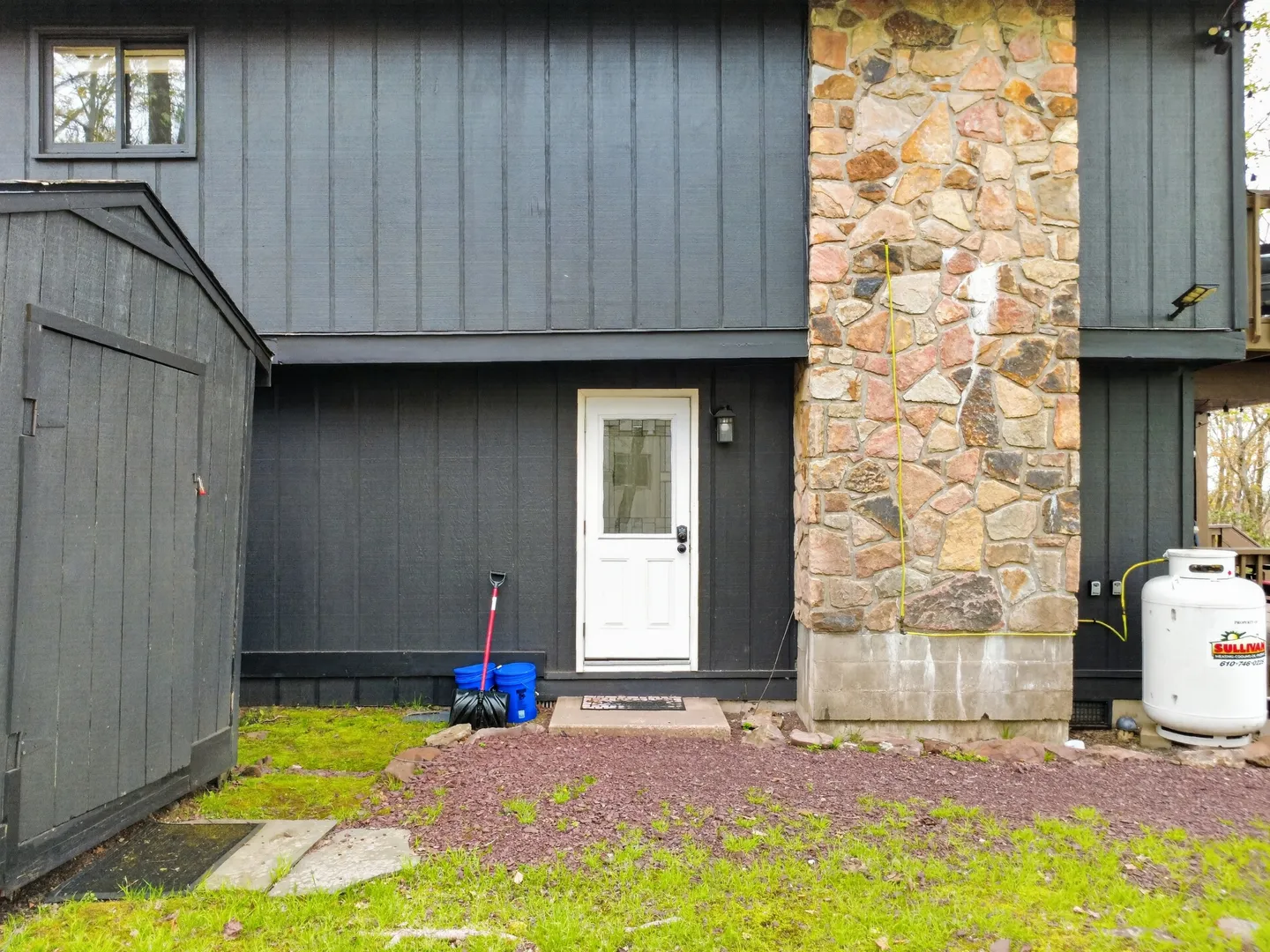 Exterior of a dark-paneled house with a white back door, stone chimney, shed, and propane tank.