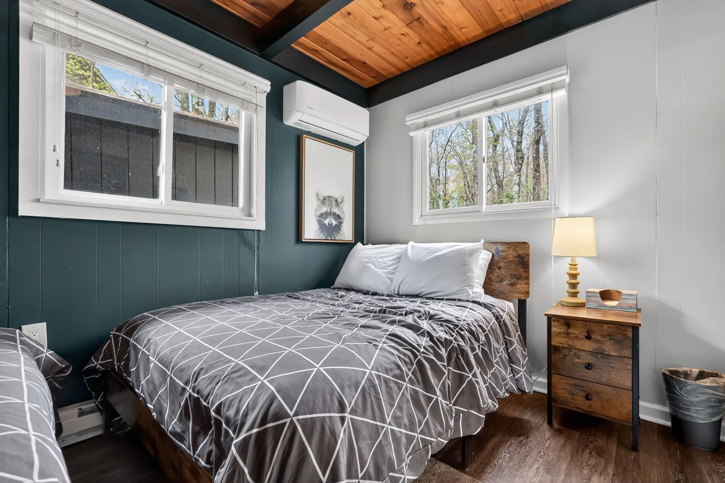 Bedroom with wood-paneled ceiling, a made bed with geometric patterned bedding, a nightstand with a lamp, and windows on adjacent walls.