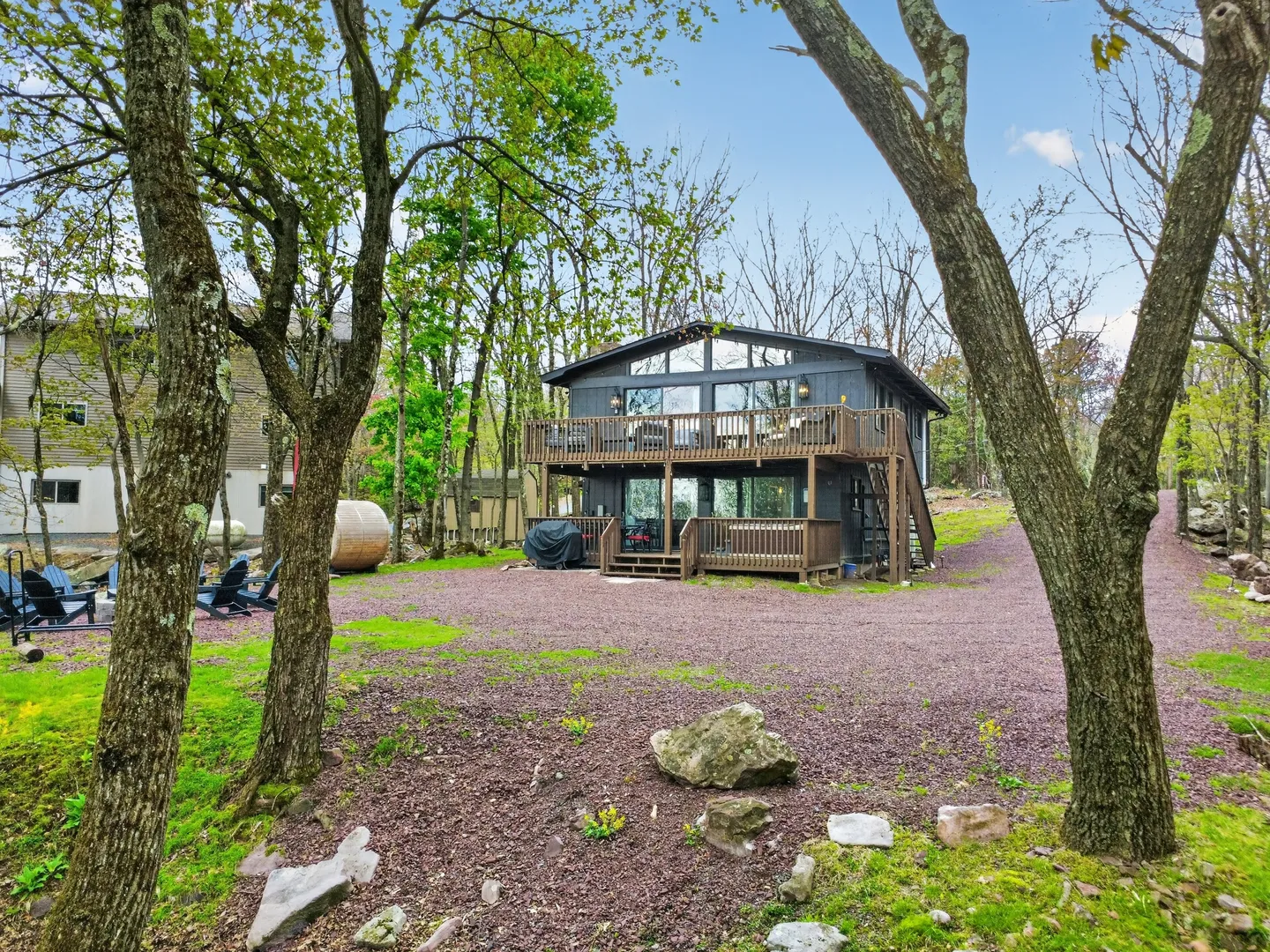 Exterior view of a dark gray two-story cabin with two levels of wooden decks, framed by trees and a gravel driveway.