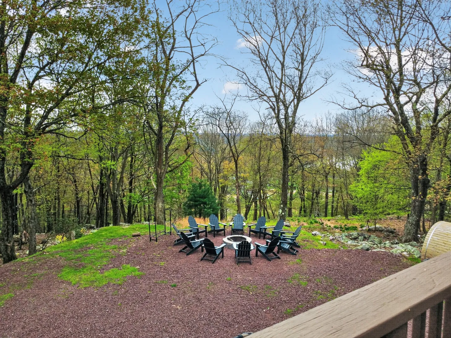 An outdoor fire pit encircled by Adirondack chairs on a gravel clearing bordered by trees.