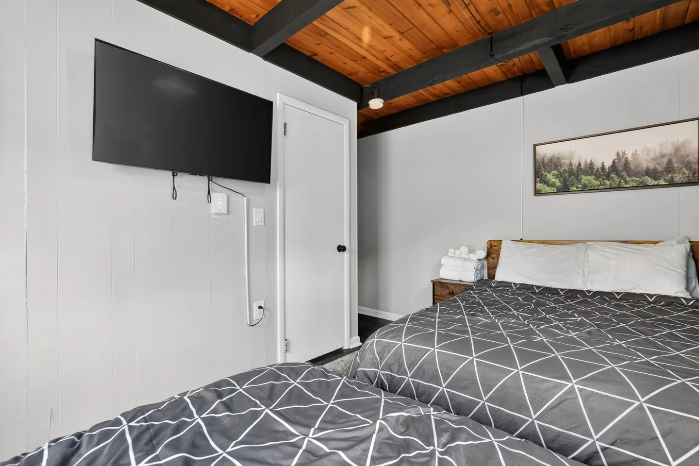 Bedroom with two beds topped with geometric patterned gray comforters, a wall-mounted TV, and wood-plank ceiling with dark exposed beams.