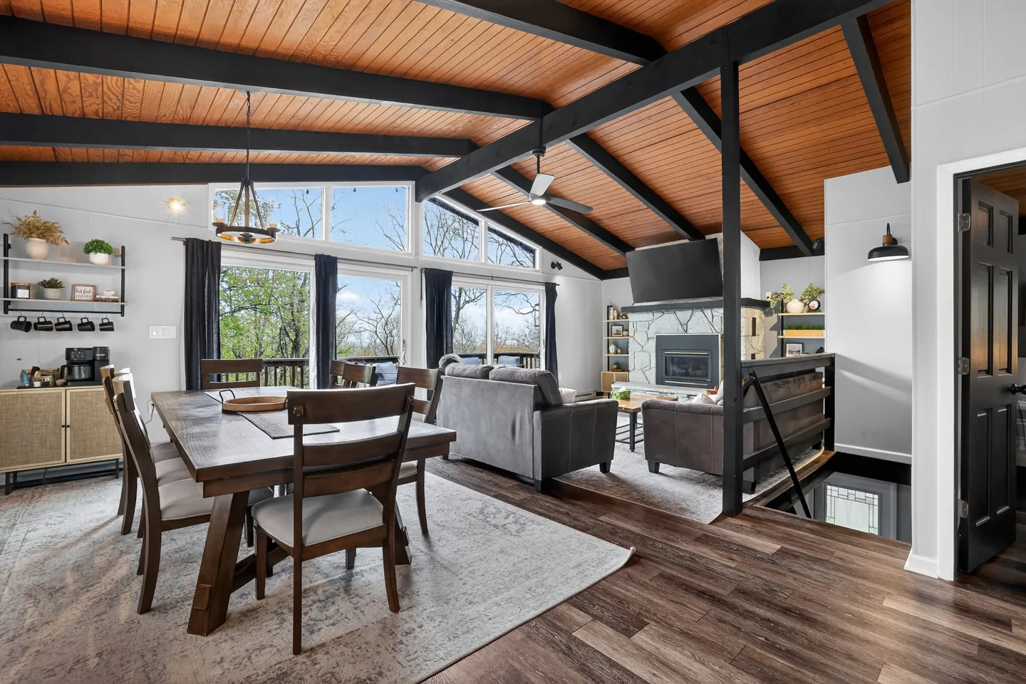 Open-concept living and dining area with wood-plank vaulted ceiling, exposed dark beams, fireplace, and large windows.
