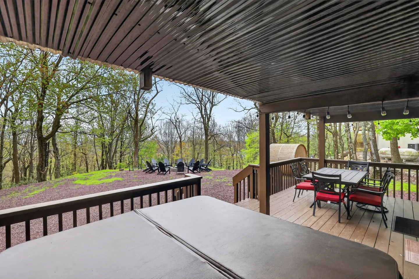 A covered deck with a hot tub cover in the foreground, outdoor dining table and chairs, and a yard featuring a fire pit surrounded by Adirondack chairs.